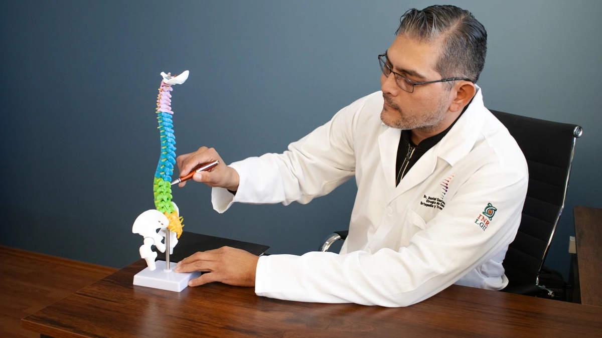 Dr. Daniel Santana López con bata blanca, explicando un modelo de columna vertebral en su consultorio